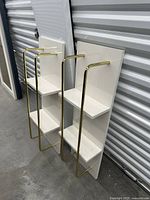 Two white wall shelves with gold metal towel bars leaning against wall