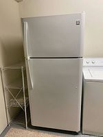 Front view of white Kenmore top-freezer refrigerator next to shelving and washer