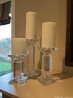 Three clear acrylic pillar candle holders of different heights with white pillar candles displayed on a shelf