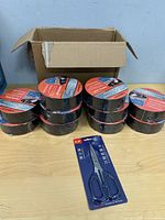 Ten shrink-wrapped tape rolls and packaged scissors beside open carton