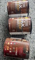 Three cans of deck stain/sealer on speckled floor—two Rust-Oleum Restore 4X Deck Coat, one Behr Premium Transparent Waterproofing Wood Finish