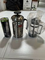 Two French presses beside beverage can; stainless steel 3M press and bubble-wrapped glass press