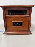 Front view of wood-finish infrared heater showing vent, control panel, remote in bag visible