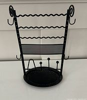 Front view of black metal jewelry stand showing earring bars, mesh panel, hooks, ring posts and base tray