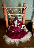 Rear view of Navajo woman doll seated at wood loom with partially woven rug, wool base visible