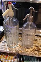 Both glass seltzer bottles with siphon tops side by side