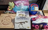 Overview of entire lot on table showing board game, Dinoworks kits, roller skates, plush toys, backpack and tin