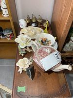 Overall lot view on wooden surface showing teapot, porcelain flowers, brass teapots, napkin rings, bowl and saucer