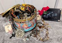 Open Fortnum & Mason carousel tin overflowing with assorted costume jewellery pieces displayed on grey surface