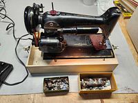 Singer black sewing machine on wooden base, yellow box of metal attachments