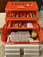 Tackle box open showing three orange trays filled with assorted lures and small clear organizer below
