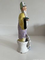 Side view of porcelain deity figurine on small white base with attached grey figure