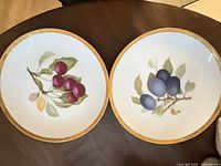 Two dessert plates showing red-purple and blue plum motifs with gold rims
