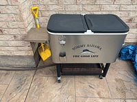 Full view of Tommy Bahama cooler on stand with side shelf and shovel nearby