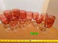 Group of 13 cranberry stemmed wine glasses on table, measuring tape visible