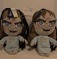 Two identical plush doll backpacks displayed front-facing showing design, straps, tags