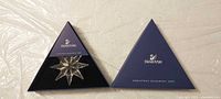 Ornament in open triangular box alongside lid displaying Swarovski branding
