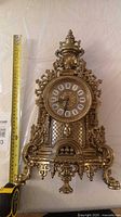 Full view of gilded mantel clock beside measuring tape showing approx 16 in height