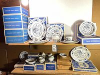 Stacked boxed dinnerware pieces on shelves showing multiple Blue Heritage pattern boxes and displayed plates