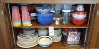 Cabinet shelf showing mixed plates, bowls, plastic cups, plastic and glass bowls, jar, boxed dish and red plastic bowl set