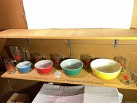 Full view of four colored Pyrex bowls, clear pitcher and four mugs arranged on shelf