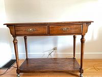 Front view of wooden serving cart showing two drawers, turned legs, shelf, casters