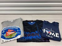 Three folded garments: grey "Come Together" tee, black skyline jersey, navy "Home" tee