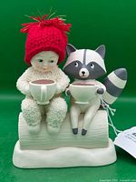 Front view of Snowbabies figurine showing Snowbaby, raccoon, mugs, red hat, log base