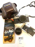 Canon AE-1 camera with brochure, leather case, Canon lens cap, Konica C35 camera visible