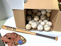 Cardboard box of about 30 baseballs with bat and glove nearby