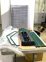 All parts laid out: green metal poles, black plastic connectors, wire shelf grid, clear plastic cover