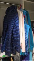 Row of outerwear hanging; navy puffer, white jacket under plastic, others obscured