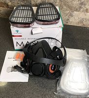 Box, respirator mask, pair of cartridge filters on top of box, sealed filter pad packs, earplugs and manual visible