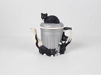 Front view of ceramic teapot showing trash can form with three black cats, lid in place