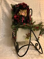 Front angled view of sleigh with doll seated, showing metal runners and greenery