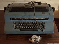 Full view of blue IBM Selectric typewriter with cord and bag of typeballs on table