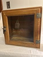 Front view of wooden display cabinet showing glass door, latch handle, hinges, and maker badge