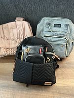 Three diaper backpacks displayed together: grey Mokaloo, black quilted, pink flap style