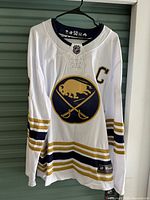Front of white Buffalo Sabres Jack Eichel jersey hanging on hanger showing logo and captain C