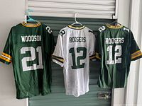 Three jerseys hung side by side: Woodson #21 green, Rodgers #12 white, Rodgers #12 green