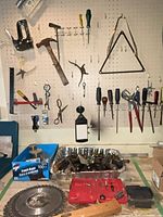 Pegboard wall with hanging hand tools: staple gun, square, hammers, pliers, screwdrivers, files; bench with drill bit cases, saw blade, tray of loose tools