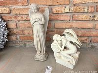Two white angel lawn ornaments against a brick wall, one standing and one sitting, showing full front view of both.