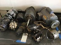 Photo showing a collection of seven electric motors of various sizes and styles, some with wiring and connectors, placed on a flat surface. Motors are visibly dirty and dusty.