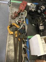 Multiple metal vices and clamps with varying sizes and one red metal pulley with hooks shown on a surface; some rust and wear visible on metal surfaces.