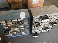 Two metal tool organizers side by side, showing multiple small labeled drawers and some contents such as bolts and screws visible in open drawers.