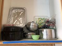 Wide view of shelf with two black speckled roasting pans, aluminum baking pan, round silver metal molds, green bowl, silverplated serving dish, and plastic floral tray
