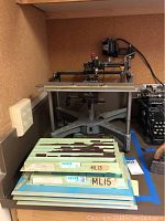 Photo of the vintage engraving machine showing its metal frame, adjustable arm, and part of the worktable along with trays of engraving plates in front.