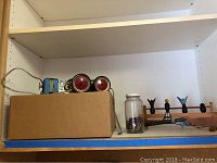 Vintage Erector set metal pieces with attached electric motor in blue and red colors stored in a cardboard box, and a glass jar containing small metal parts next to a wooden target setup.