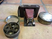 Kodak TBI folding camera with pink bellows, lens, and metal body. Two adjacent round metal film cans, one open with film spools inside.