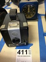 Kodak Brownie Hawkeye camera with black and silver body and carrying strap, placed next to a blue interval timer with a clock face on it
